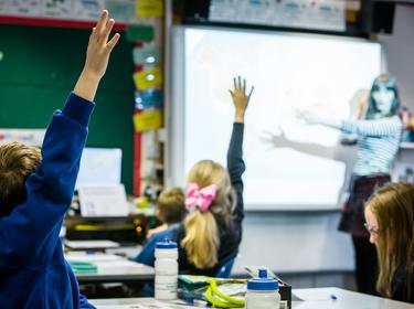 Children raising their hands in a classroom while a teacher points to the board